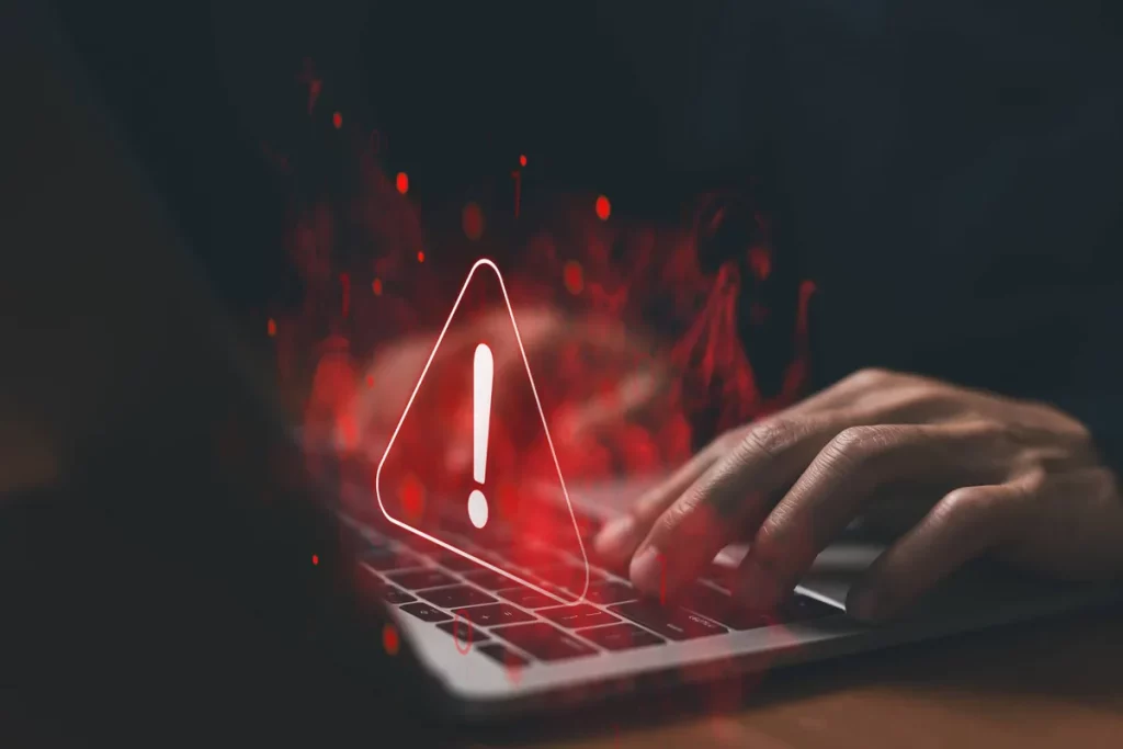 Um usuário digita em laptop com símbolo de alerta e efeitos digitais vermelhos, ilustrando ameaça cibernética. (Foto: olhardi