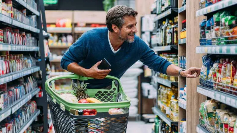 Homem escolhe produtos em supermercado, com carrinho de compras e celular na mão. (Foto: DECO.PROTESTE.PT)