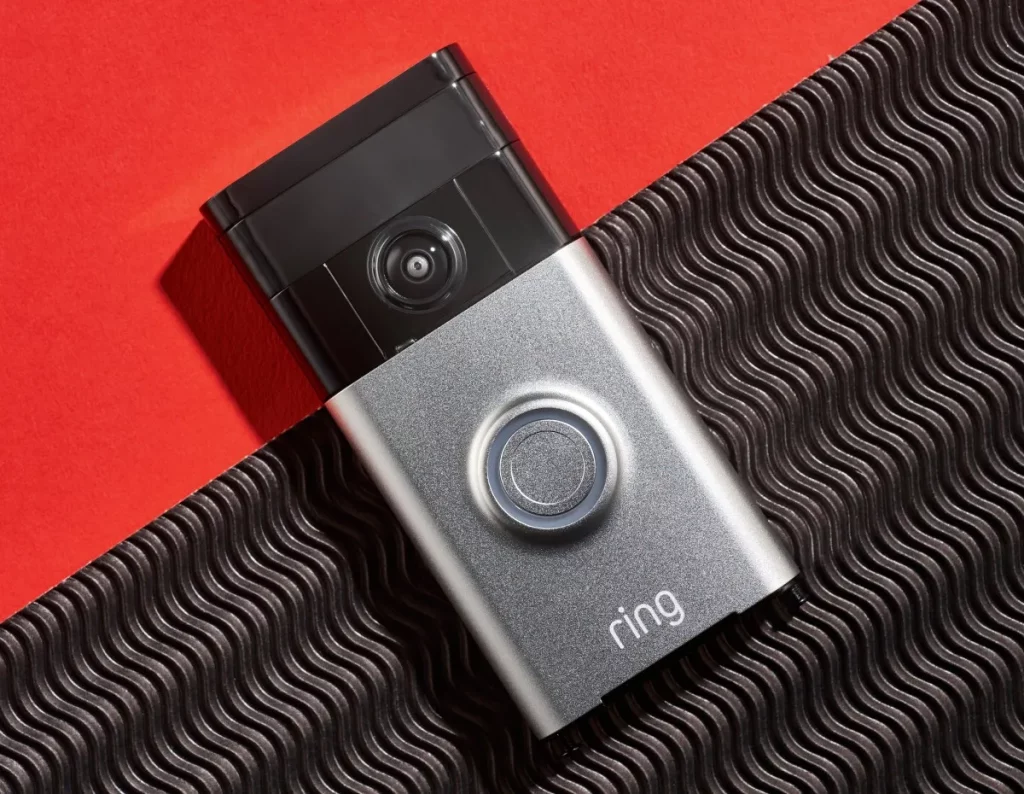 Câmera de porta inteligente da Ring em destaque sobre fundo vermelho e textura ondulada. (Foto: techcrunch.com)