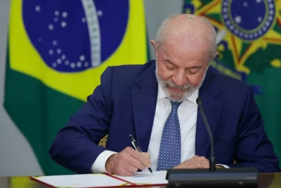 O presidente Lula assina documento oficial em cerimônia com bandeira do Brasil ao fundo. (Foto: www.metropoles.com)