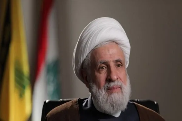 Sheikh Naim Qassem, líder do Hezbollah, em foto oficial com bandeiras do movimento ao fundo. (Foto: en.mehrnews.com)