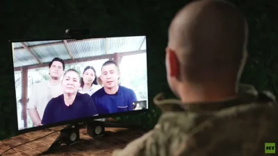 Um homem em uniforme assiste a uma videochamada com sua família, que aparece na tela. (Foto: rt.com)