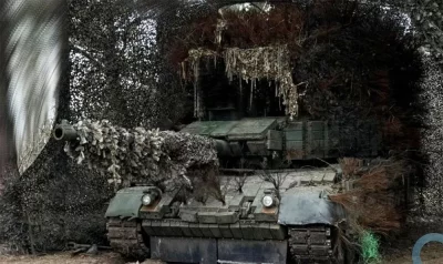 Tanque blindado camuflado em posição de combate, com vegetação e telas de camuflagem. (Foto: defesanet.com.br)