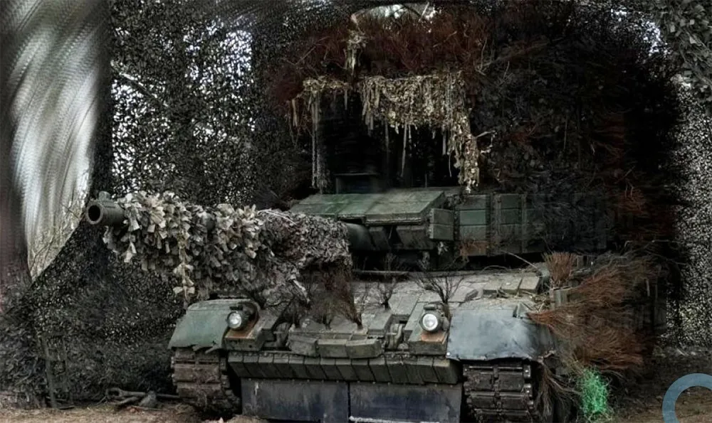 Tanque blindado camuflado em posição de combate, com vegetação e telas de camuflagem. (Foto: defesanet.com.br)